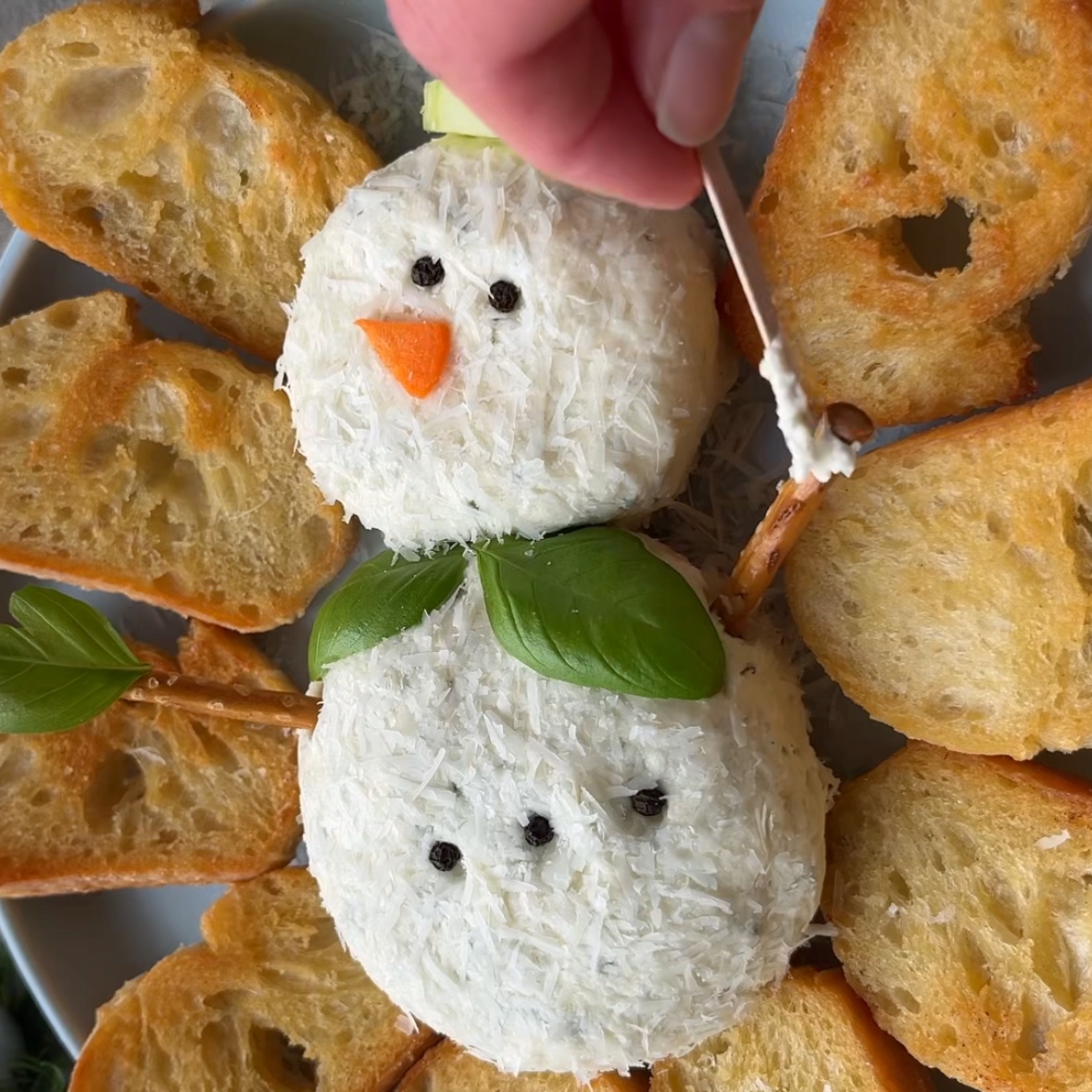 A hand adds the final details to a festive Christmas Cheese Ball shaped like a snowman, with bread slices surrounding it.