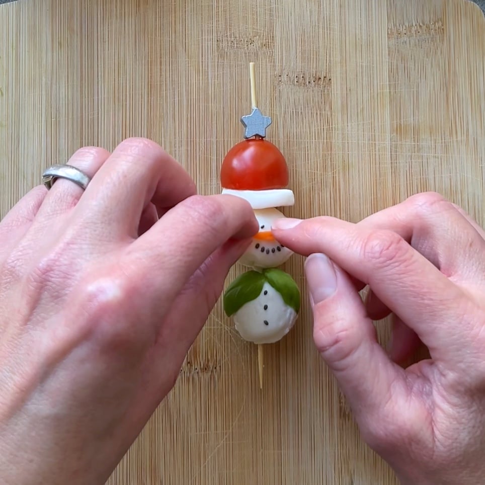 Two hands assemble a snowman-shaped snack on a wooden board using a skewer with tomato, mozzarella, and basil.