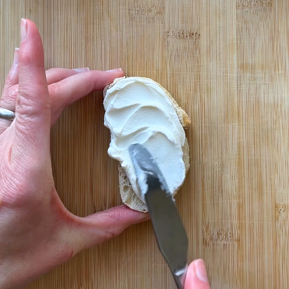 A hand spreads cream cheese on a slice of bread using a butter knife.