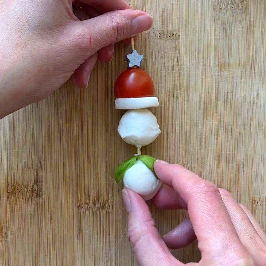 Hands assembling a skewer with a cherry tomato, mozzarella balls, a basil leaf, and a slice of mozzarella cheese on a wooden cutting board.