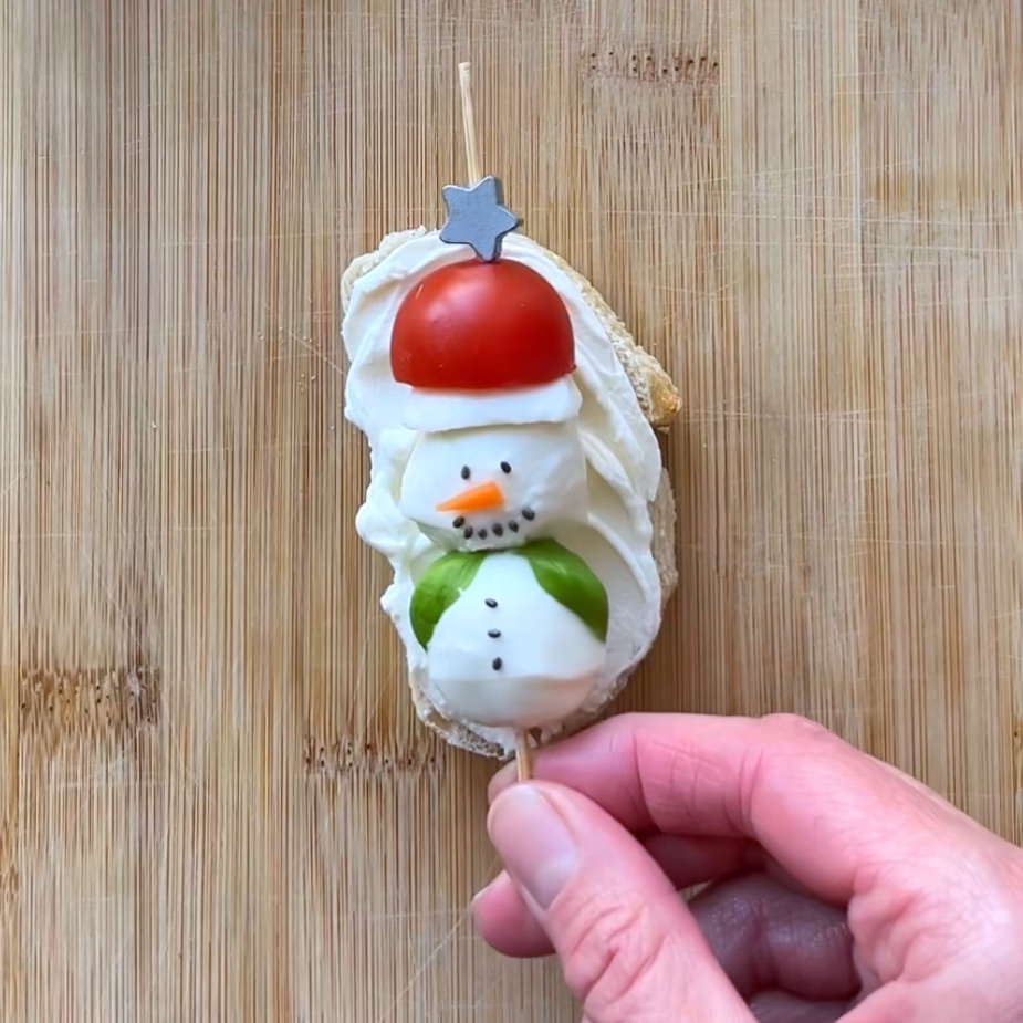 A hand holds a skewer with a snowman made of mozzarella, tomato, and basil on cream cheese&ndash;spread bread.