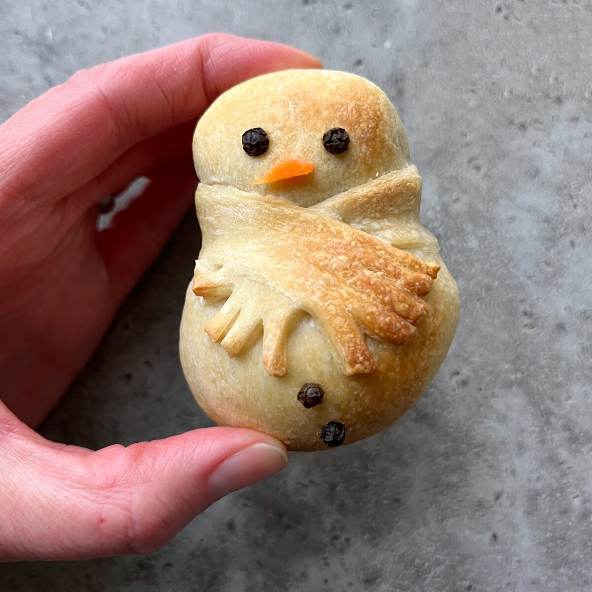 A hand holds a small sausage roll shaped like a snowman, decorated with peppercorns for eyes and buttons, and a piece of dough for a scarf.