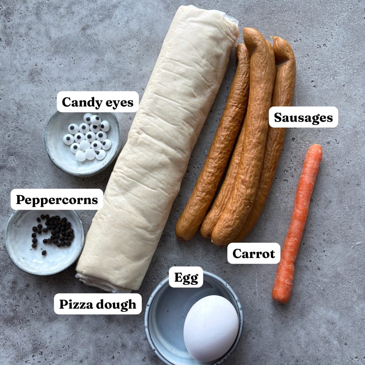 Pizza dough, sausages, a carrot, an egg, peppercorns, and candy eyes are arranged on a gray surface, each item labeled as the essential ingredients for making festive Christmas Sausage Rolls.