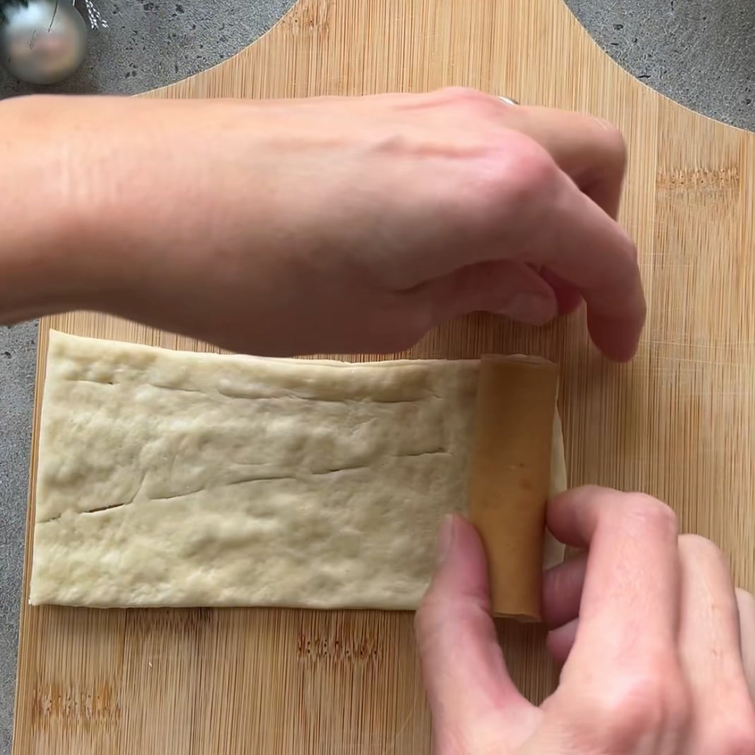 Hands rolling a sausage into a sheet of dough on a wooden cutting board.