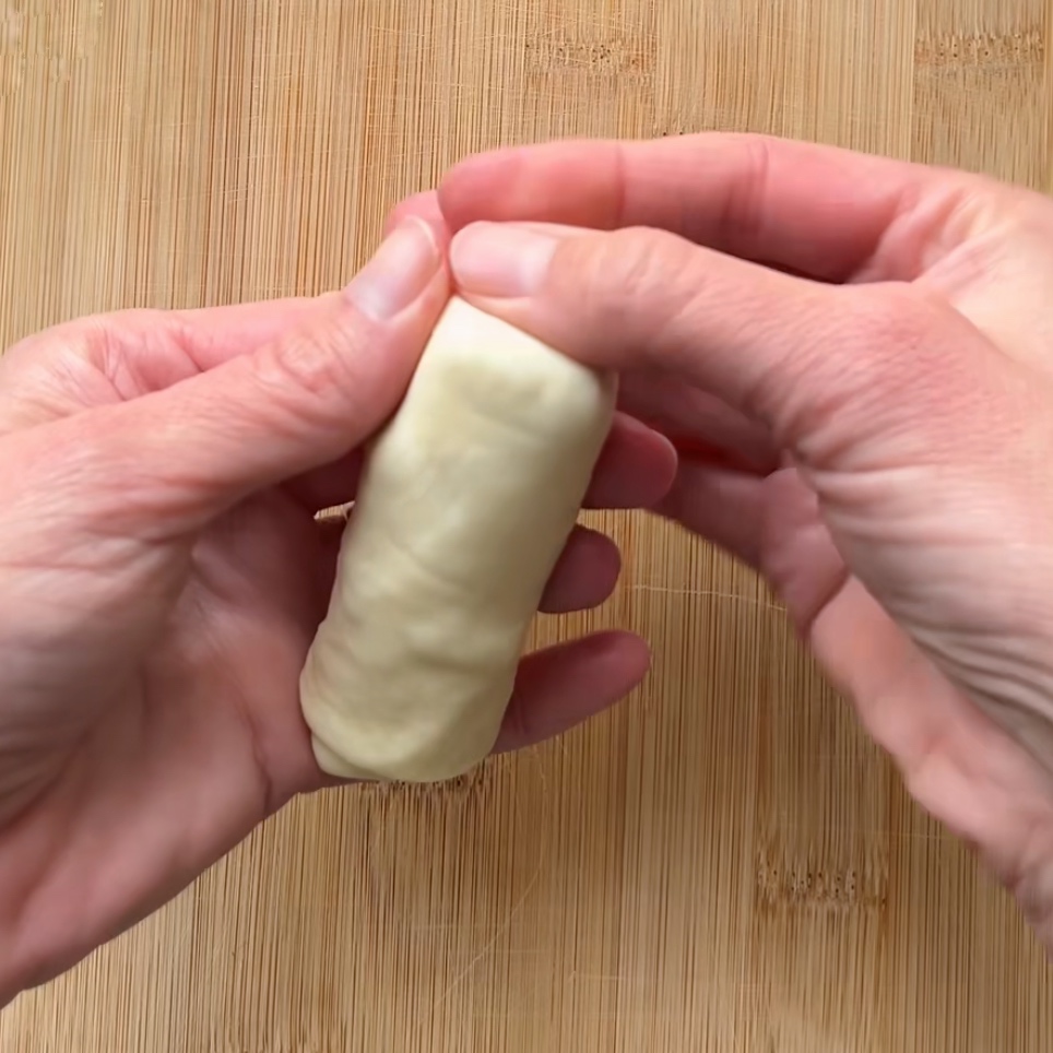 Two hands carefully shaping a small roll of dough on a wooden surface.