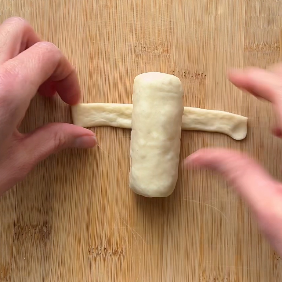 Two hands wrap a strip of dough around a cylindrical piece of dough on a wooden surface.