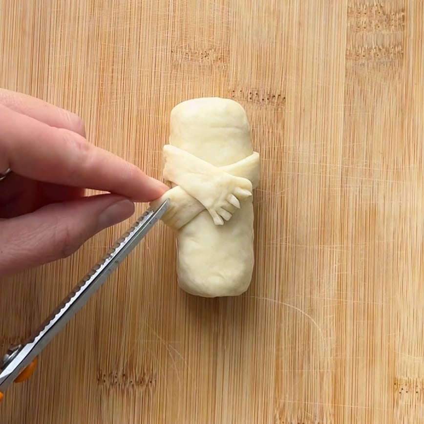 A hand uses a knife to cut fringe on crossed strips of dough wrapped around a small Christmas Sausage Roll, on a wooden cutting board.