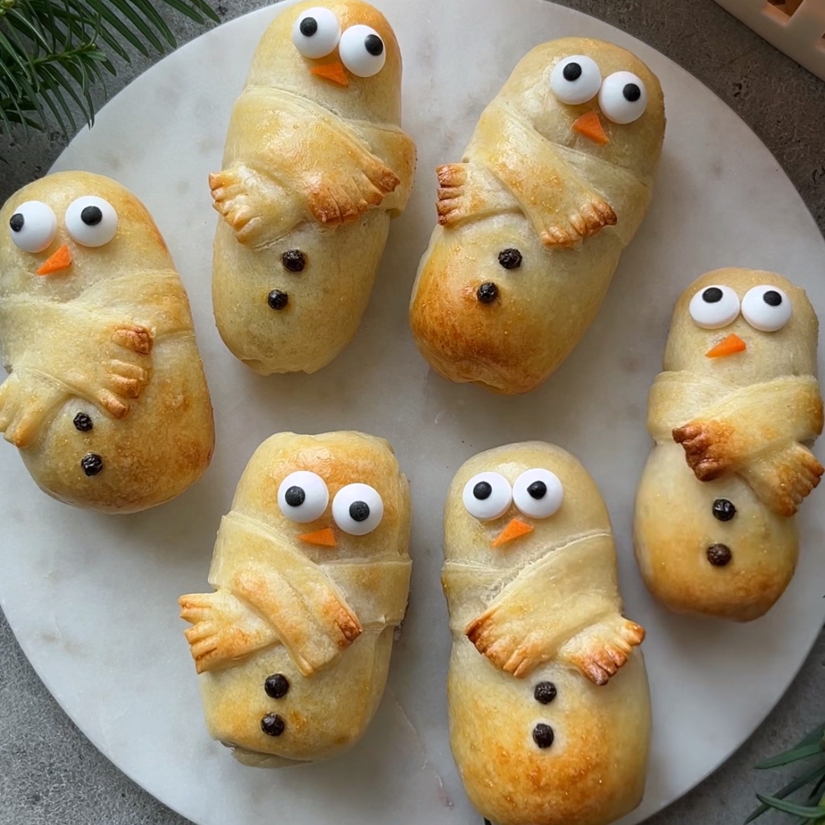 Six sausage rolls decorated as snowmen with large candy eyes, orange noses, and peppercorn buttons, arranged on a round white surface.