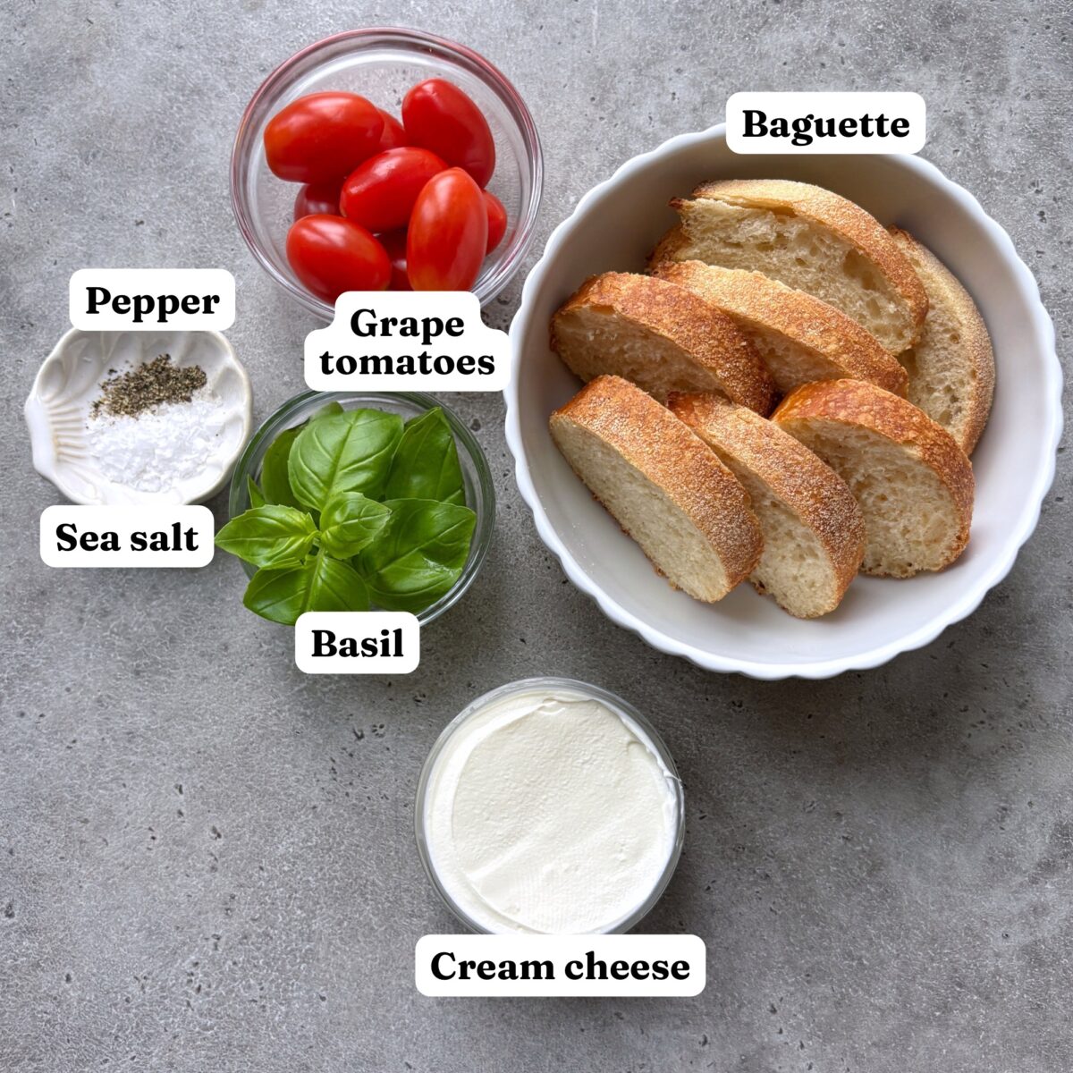 Sliced baguette surrounded by bowls of grape tomatoes, fresh basil, cream cheese, sea salt and pepper.