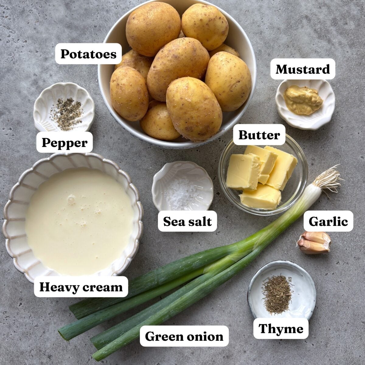 Labeled ingredients on a gray surface for delicious Potato Stacks: potatoes, mustard, pepper, sea salt, butter, garlic, thyme, heavy cream, and green onions.