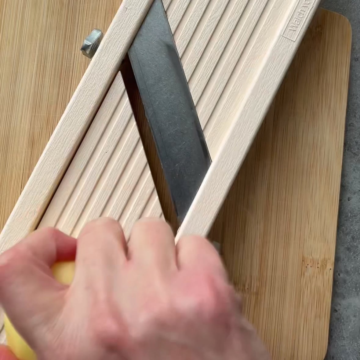 A hand slices a potato using a mandoline slicer on a bamboo cutting board.