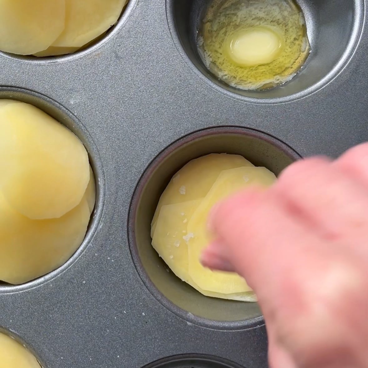 A hand sprinkles salt on sliced potatoes placed in a muffin tin.
