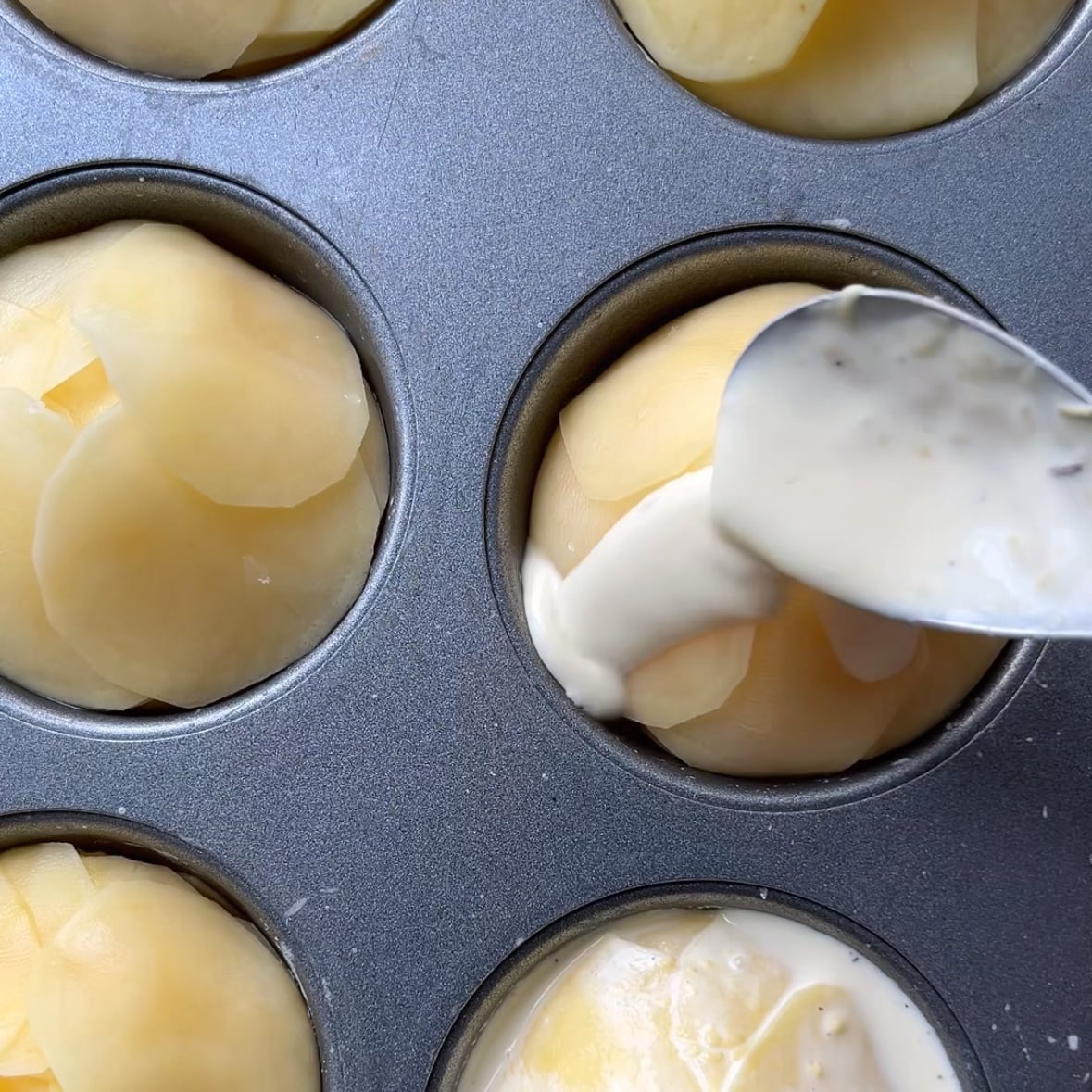 A spoon pours creamy sauce over thinly sliced potatoes arranged in a muffin tin.