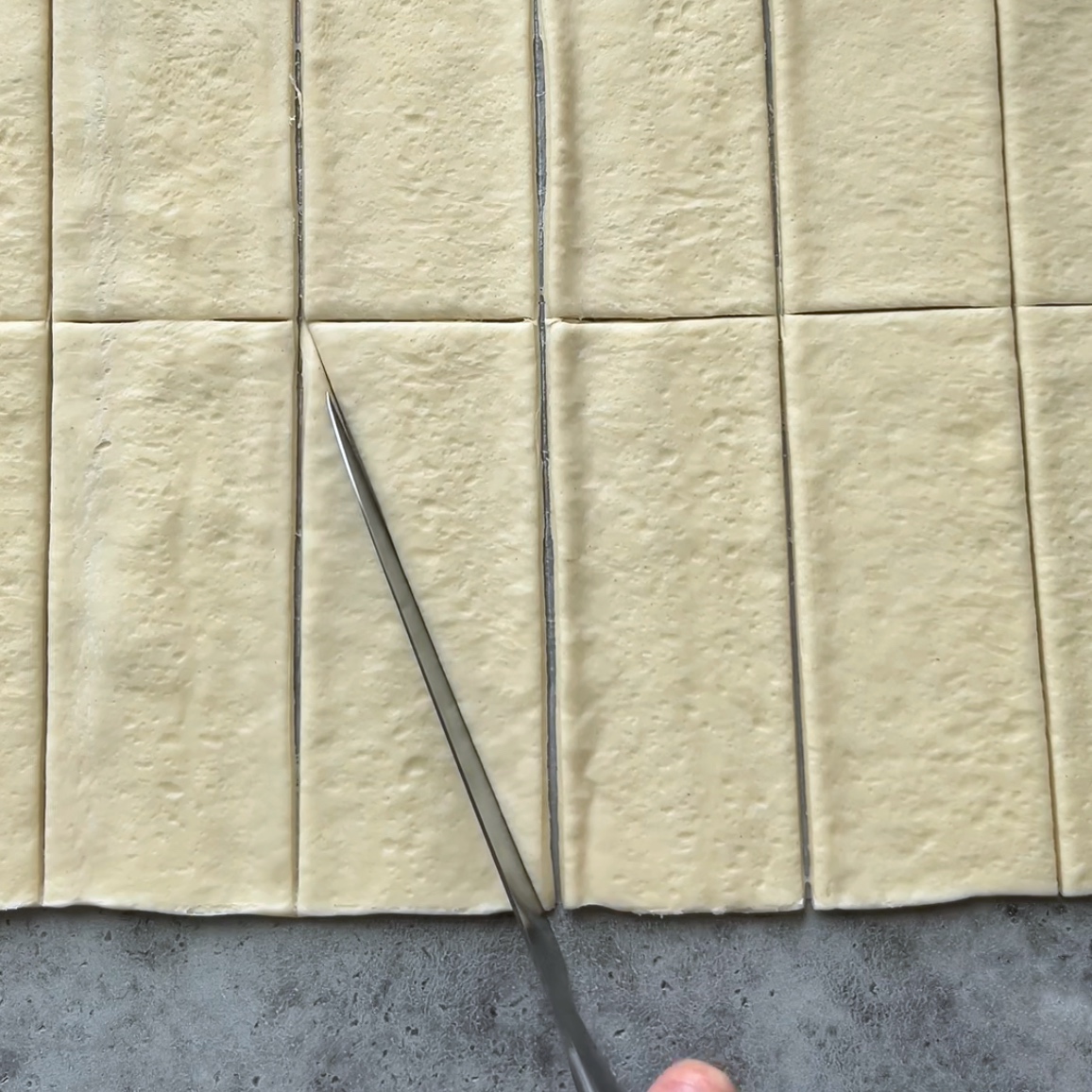 A knife is cutting a sheet of dough into evenly sized rectangular pieces on a gray surface.