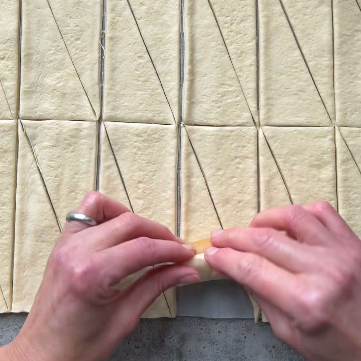 Hands rolling a sausage into a triangle of dough, surrounded by other triangles cut from a large sheet.
