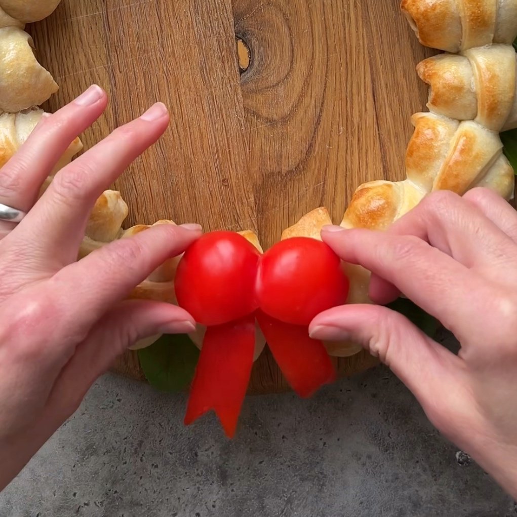 Two hands position a red pepper bow as a bow decoration on a sausage roll wreath.