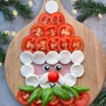 A festive Smiling Santa Caprese face made from tomato slices, mozzarella rounds, ham, basil leaves, and a cherry tomato nose arranged on a wooden board with pine branches and string lights in the background.