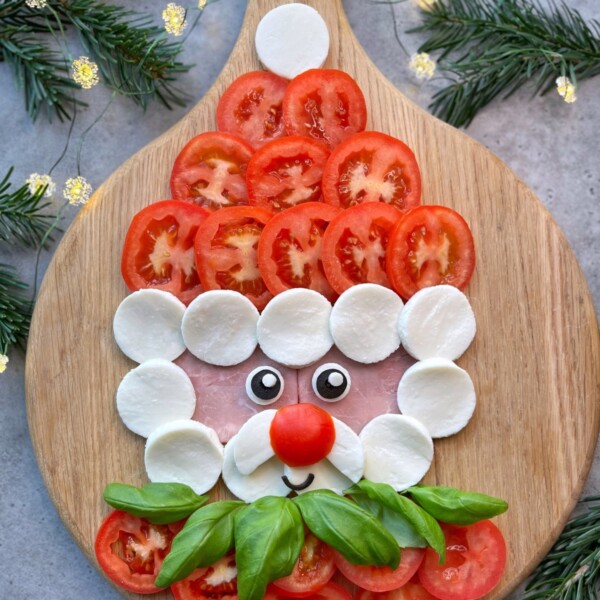 A festive Smiling Santa Caprese face made from tomato slices, mozzarella rounds, ham, basil leaves, and a cherry tomato nose arranged on a wooden board with pine branches and string lights in the background.