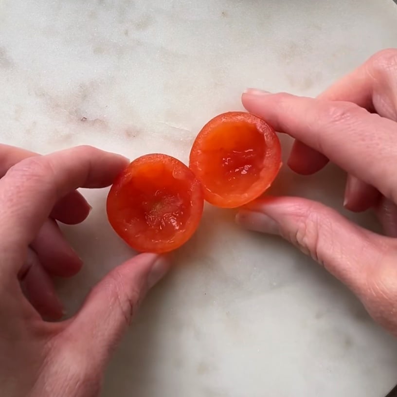 Two hands hold a cherry tomato cut into halves with the flesh removed.