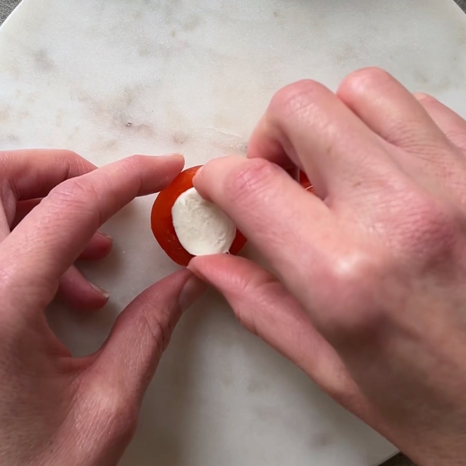 A hand places a bocconcini half into a halved tomato.