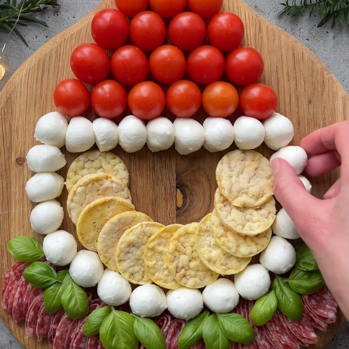 A wooden board holds a festive Christmas Charcuterie Santa face made of cherry tomatoes, mozzarella balls, rice crackers, basil leaves, and salami, with a hand placing a mozzarella ball.