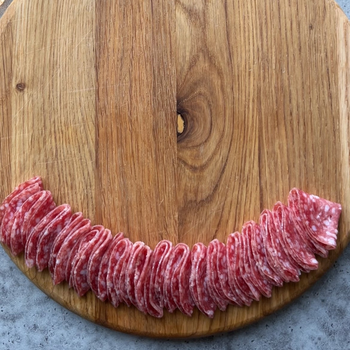 Slices of salami arranged in a curved line on a round wooden cutting board, placed on a gray surface.