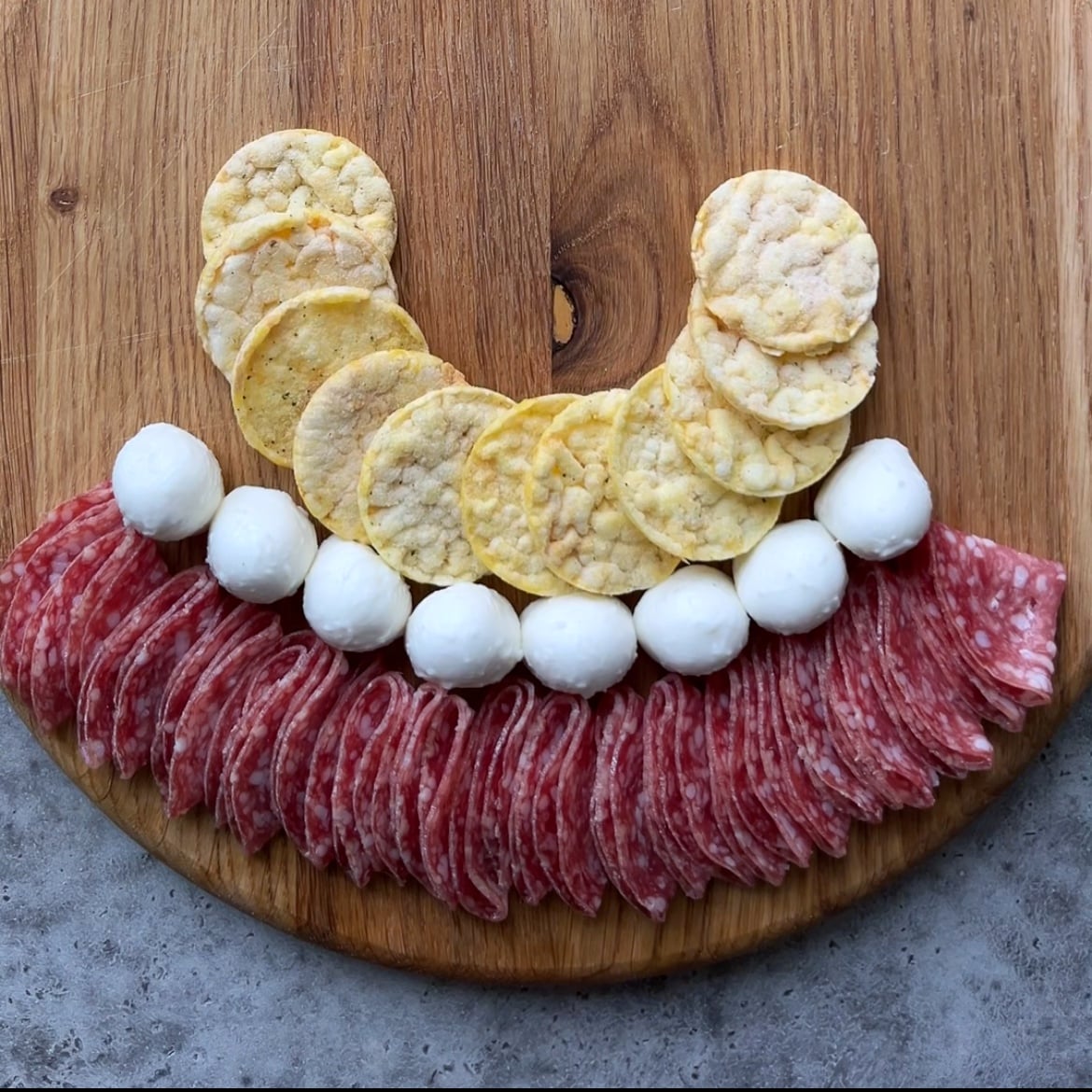 A wooden board with rows of salami slices, mozzarella balls, and round rice crackers artfully arranged in a semicircle.