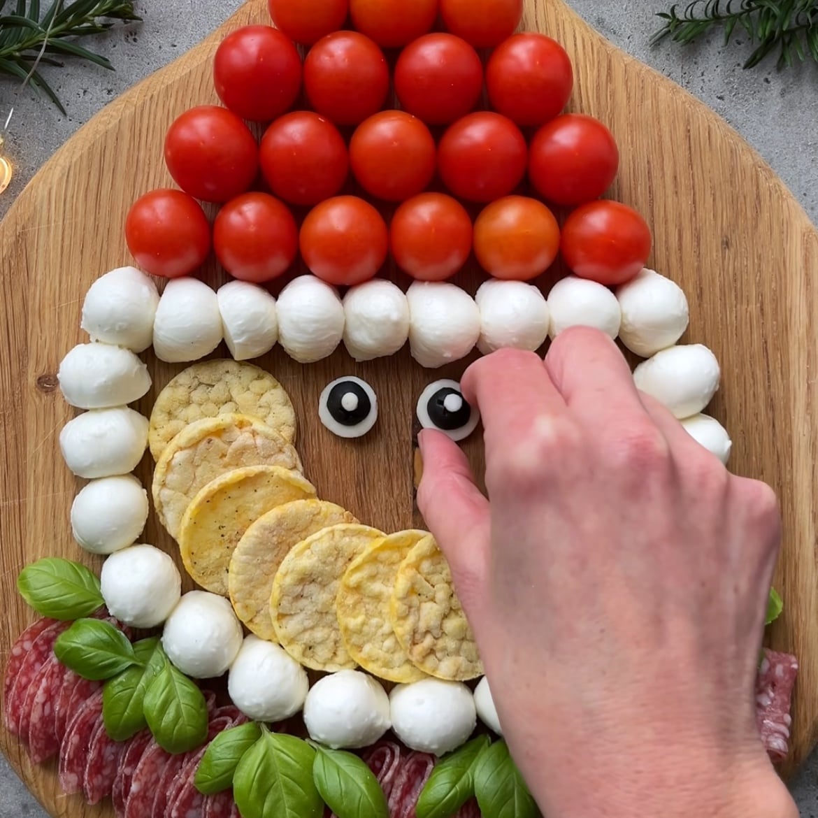 A hand arranges olive slices on mini mozzarella slices to create eyes on a Santa face made of tomatoes, mozzarella, crackers, basil, and salami.