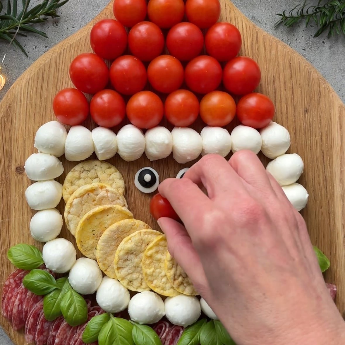 A hand places a tomato on a platter arranged with tomatoes, mozzarella, crackers, basil, and salami to resemble a Santa face.