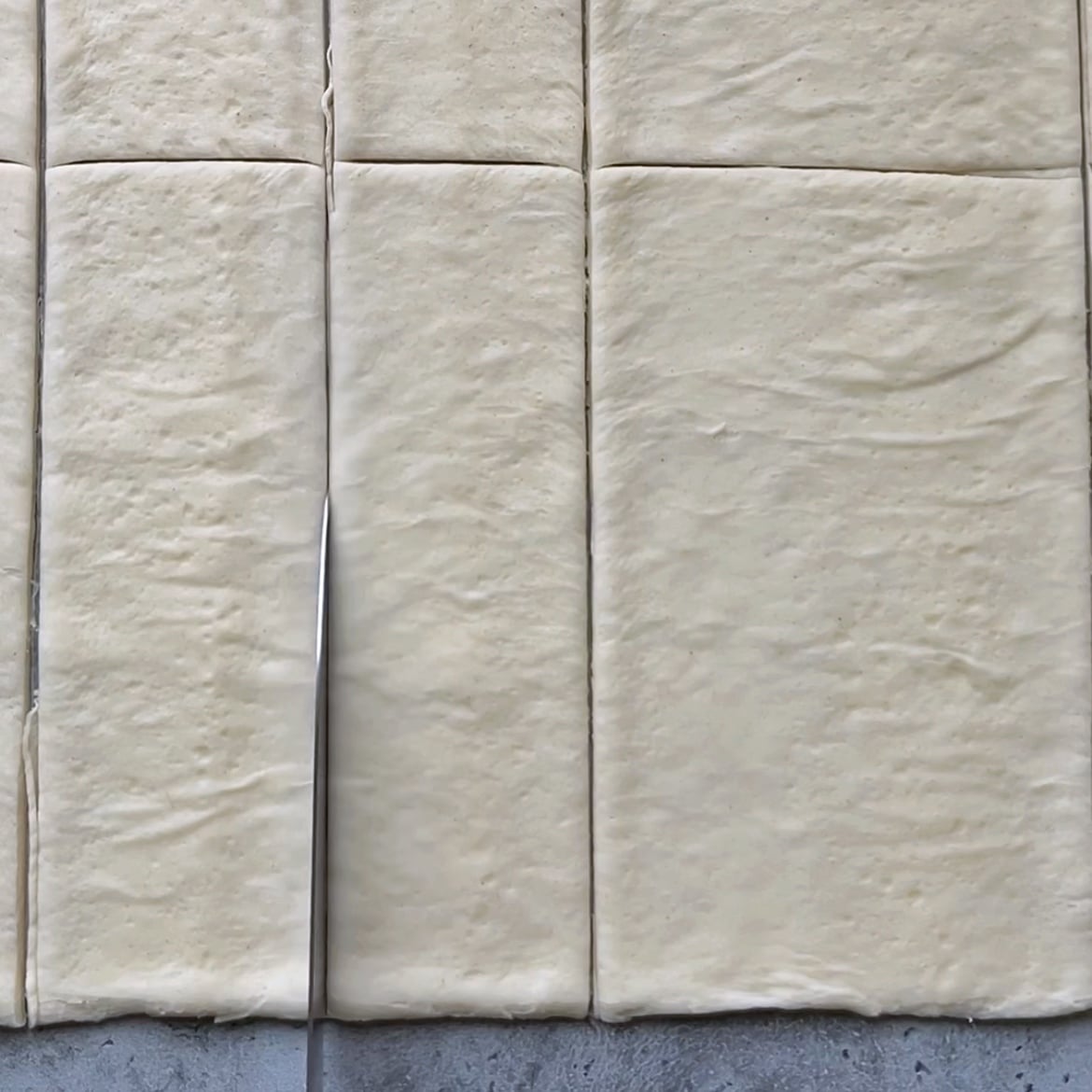 A knife cuts rectangular pizza dough into symmetrical rectangular pieces.