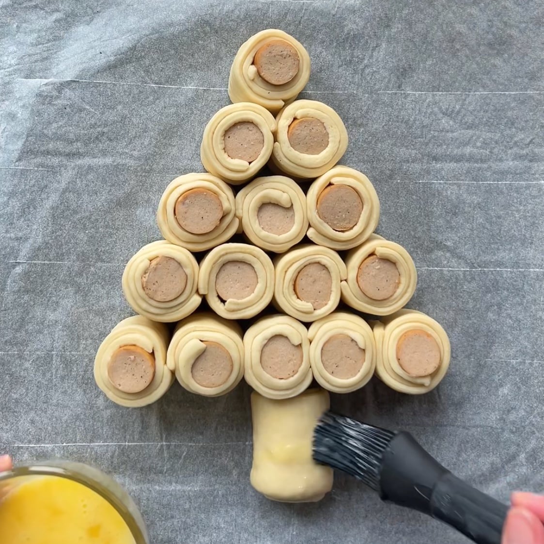 A hand brushes egg wash onto sausage rolls arranged in the shape of a Christmas tree.
