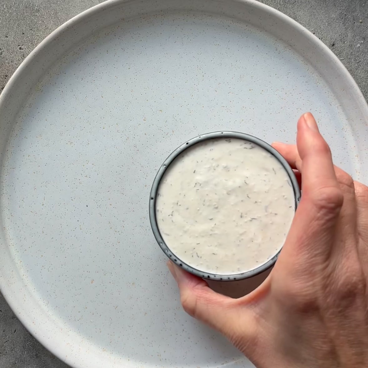 A hand holds a small bowl filled with a creamy white dip over an empty white plate on a gray surface.