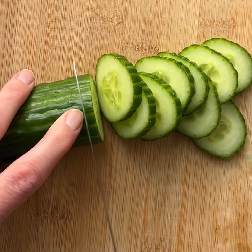 A hand is slicing a cucumber on a wooden cutting board with a kitchen knife.