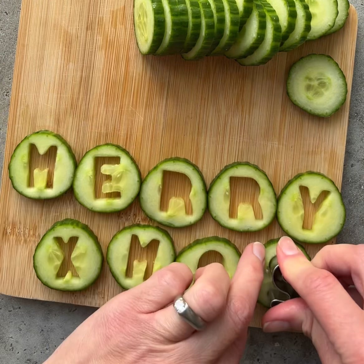 Sliced cucumbers on a wooden board with letters cut out to spell MERRY XMAS. A personโs hands cut the last letter.