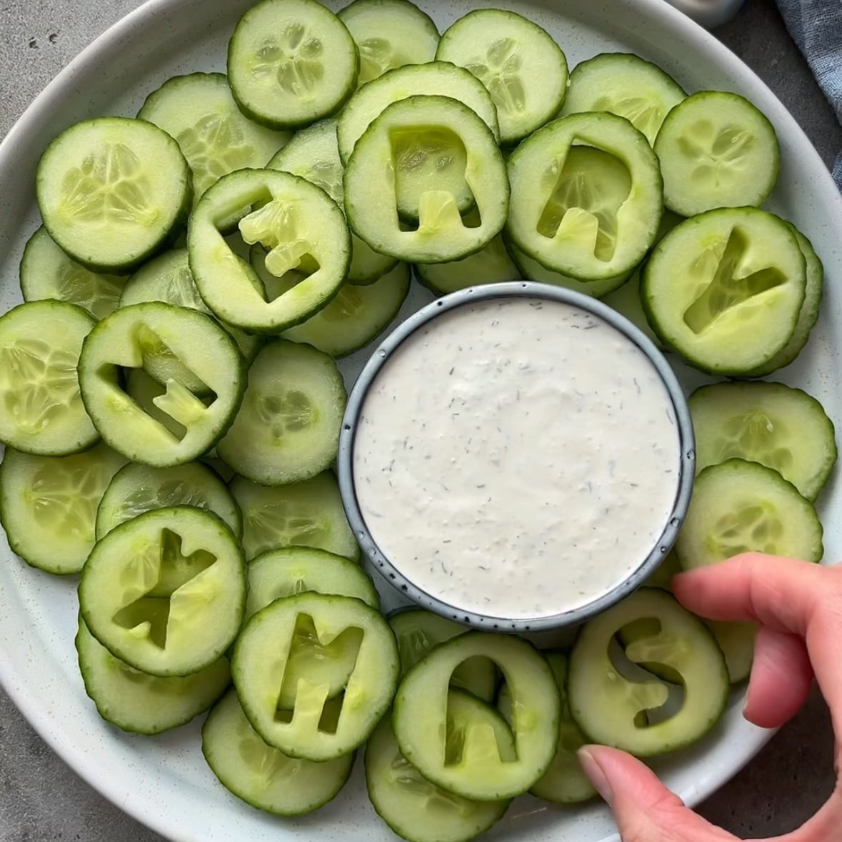 A festive Christmas Veggie Tray features a plate of cucumber slices spelling MERRY XMAS around a bowl of dip, while a hand arranges a slice.