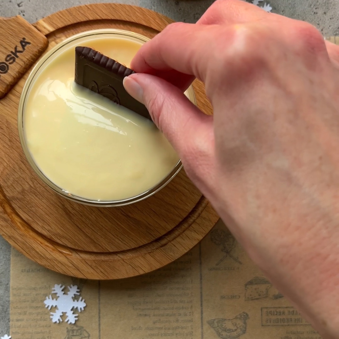 A hand dips a chocolate cookie into a glass bowl filled with creamy white chocolate, placed on a round wooden coaster.