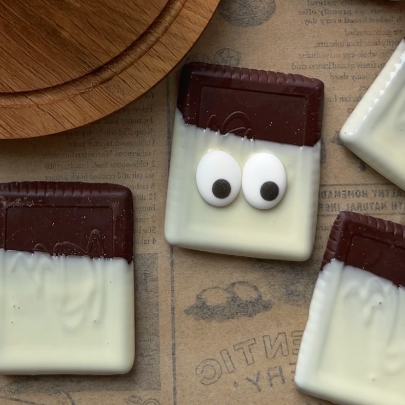 A rectangular cookie dipped in white chocolate with candy eyes is among other similar cookies on parchment paper.