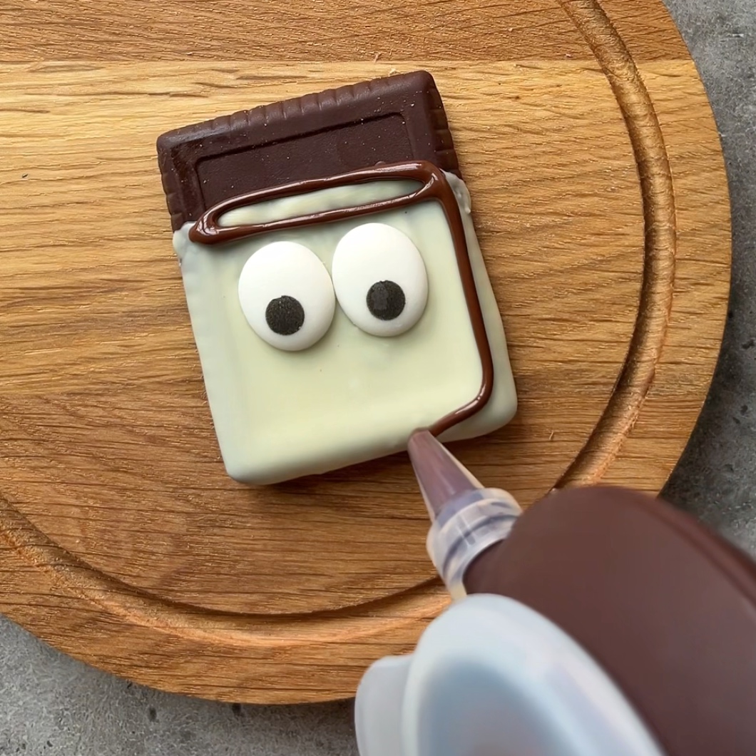 A hand decorates a rectangular, chocolate-covered cookie with googly eyes.