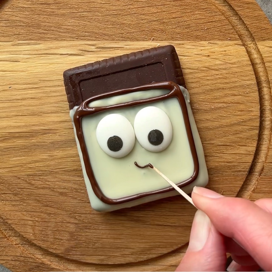 A hand uses a toothpick to draw a smile with chocolate on a cookie decorated with white chocolate and large candy eyes.