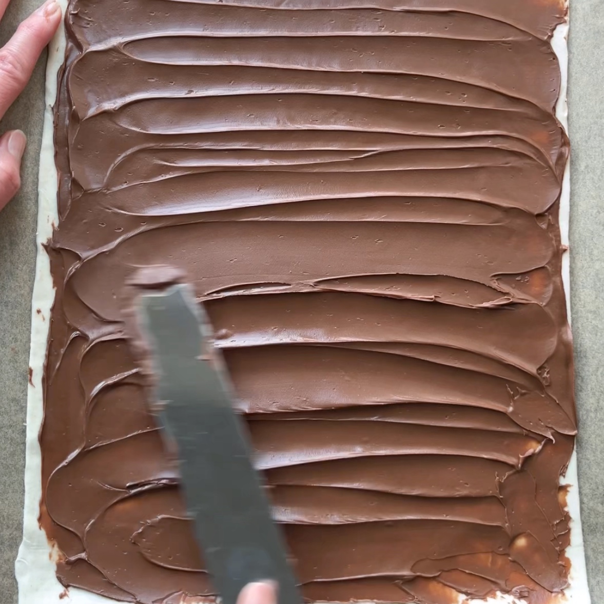 A person spreads chocolate hazelnut spread over a rectangular sheet of pastry dough with an offset spatula.