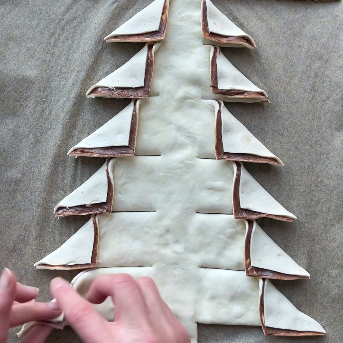 Hands folding pastry dough slices shaped like a Nutella Christmas Tree on parchment paper, with chocolate spread visible between layers.
