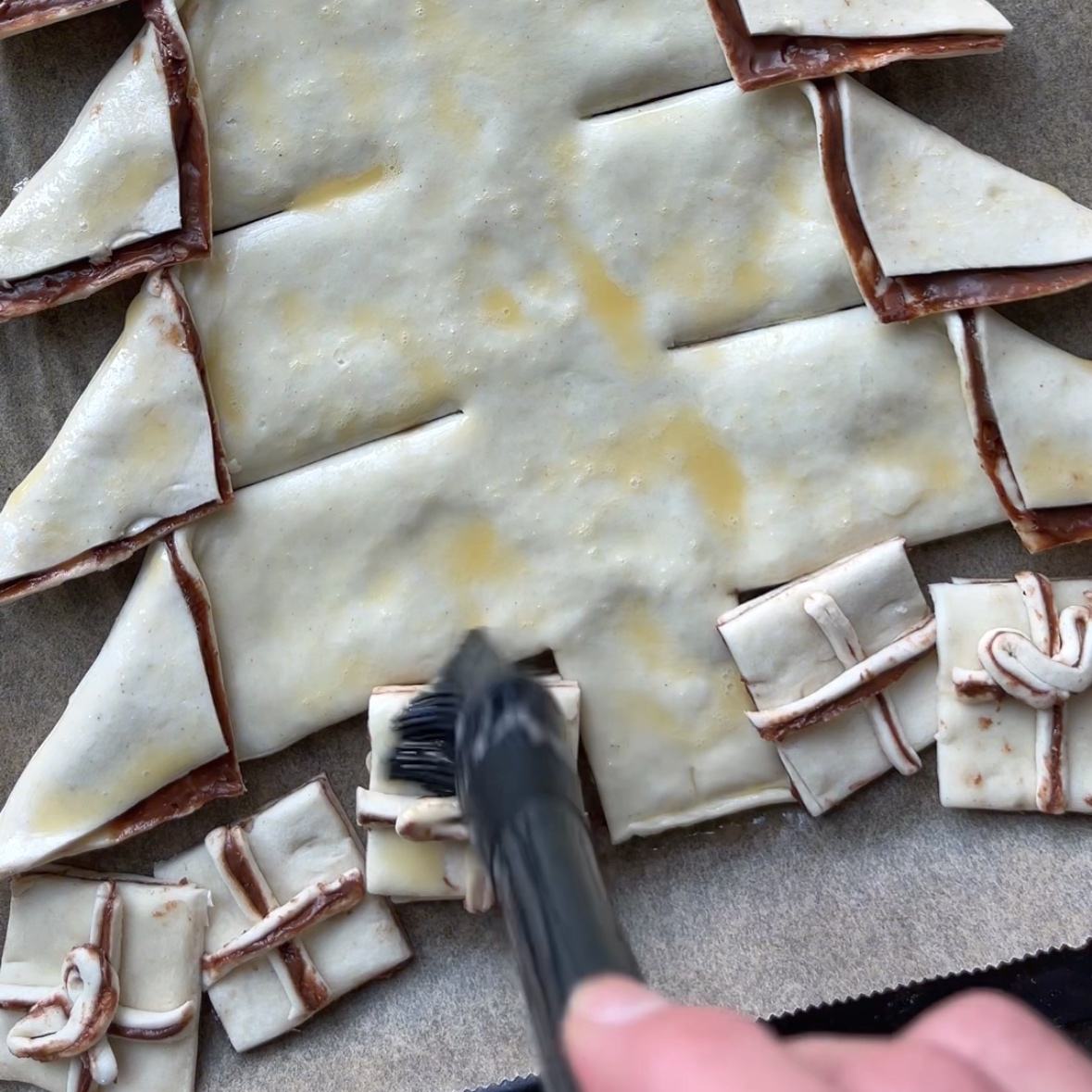 A hand uses a brush to apply egg wash to pastry dough shaped like a Nutella Christmas Tree, with small present-shaped pastries at its base.