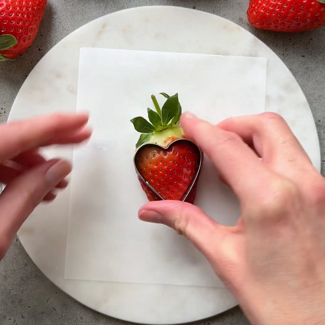 A hand uses a cookie cutter to cut out a heart from of a strawberry half.