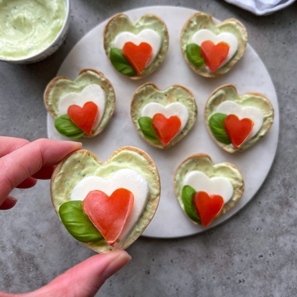 Heart-shaped Caprese Bites with green avocado spread, fresh basil, a mozzarella heart, and a tomato heart on top.
