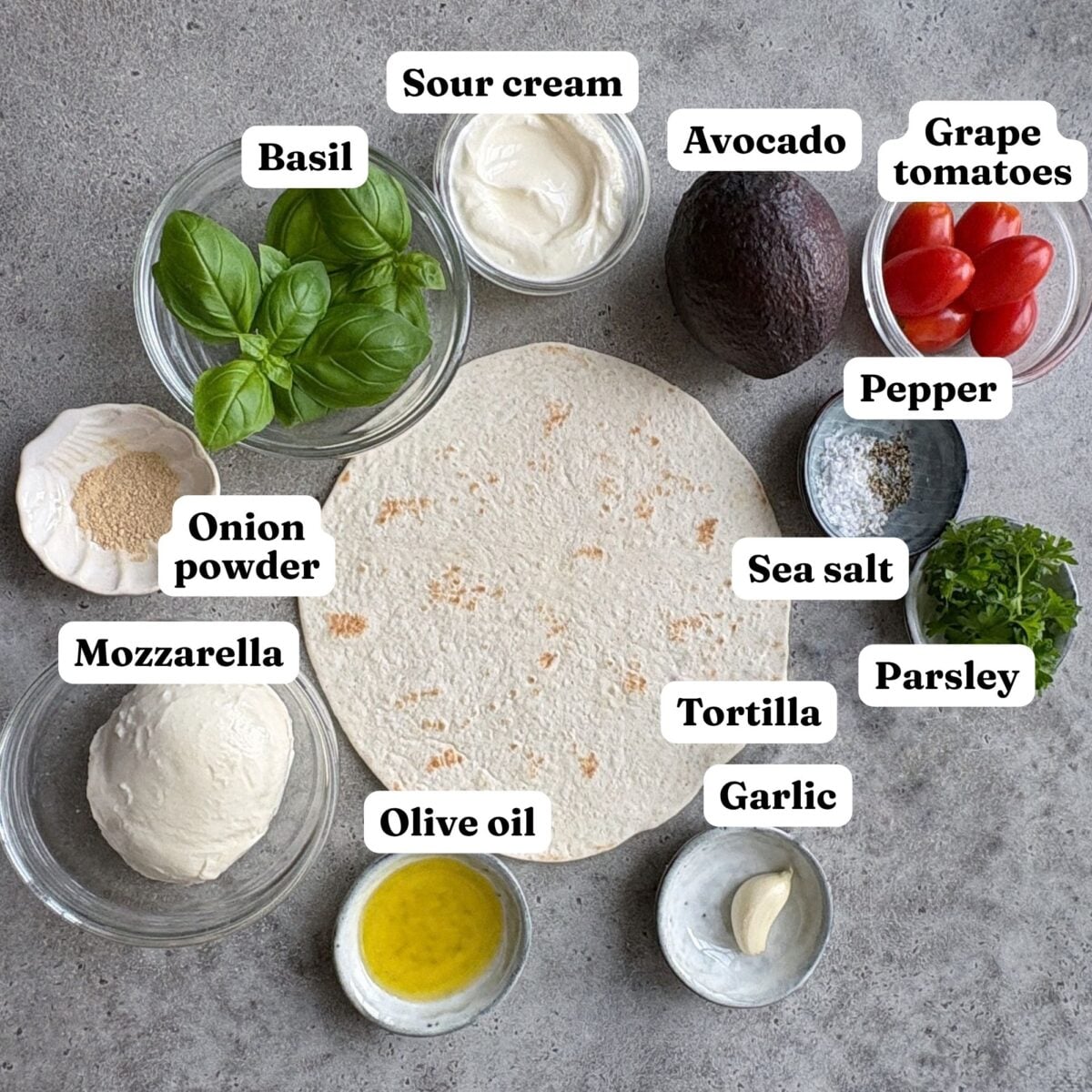A flat tortilla surrounded by ingredients including avocado, grape tomatoes, mozzarella, olive oil, and garlic.