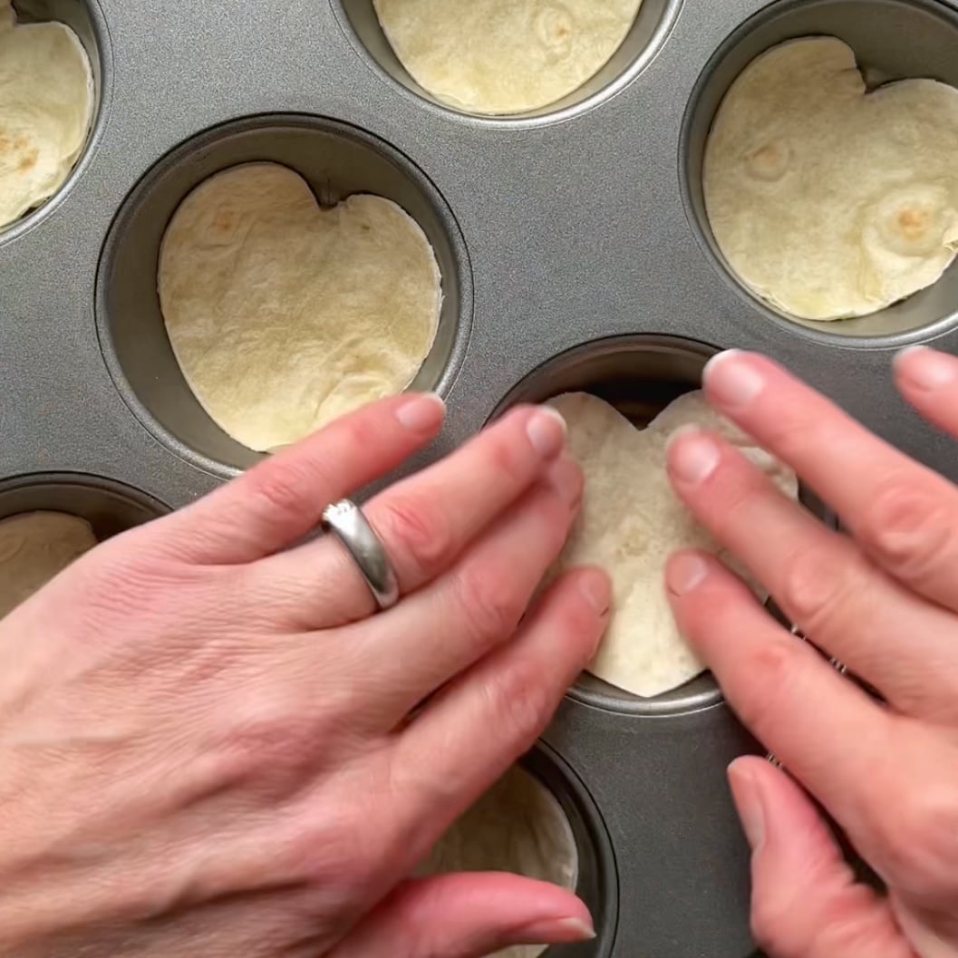 Two hands push a tortilla heart cut out into a muffin tin cavity.
