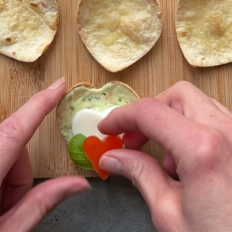 A hand tops a small tomato heart onto a tortilla heart-shaped shell topped with avocado cream, fresh basil, and a mozzarella heart.