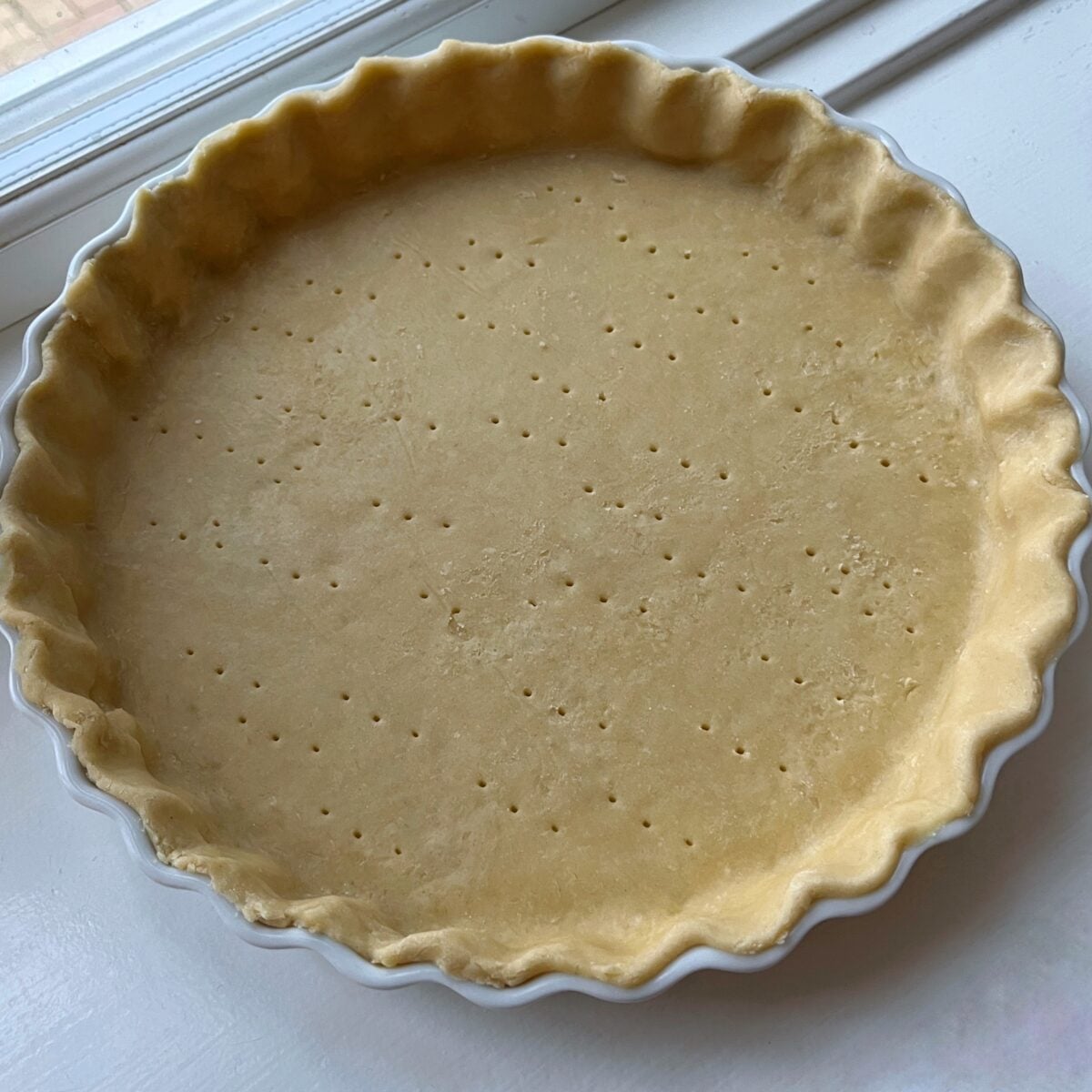 Unbaked pie crust in a fluted ceramic dish, with fork holes evenly pricked across the surface, sitting on a windowsill.