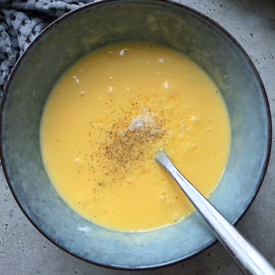 A bowl of beaten eggs with black pepper and salt, ready to become the base for a delicious Chicken Quiche, with a spoon resting inside the bowl on a light countertop.