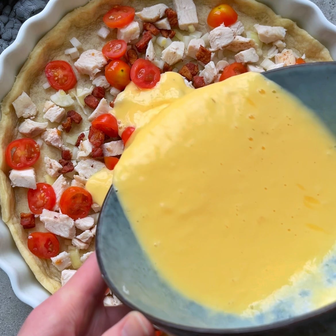 A hand pours a yellow egg mixture over diced chicken, tomatoes, onions, and bacon pieces in a pie crust.